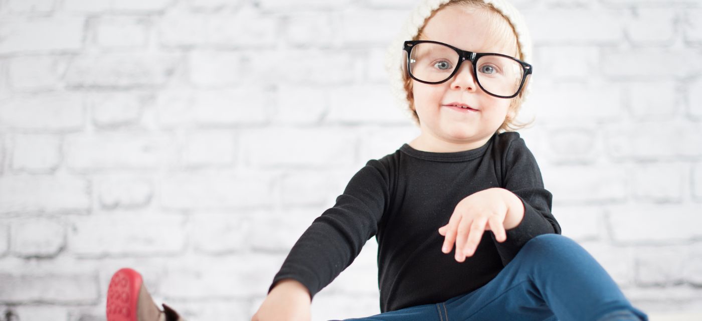 Toddler wearing dark rimmed glasses sitting in front of brick wall.